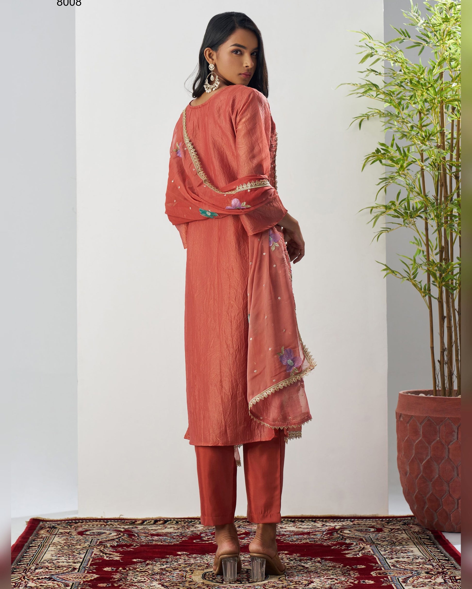 Woman in a peach-colored traditional outfit standing in a room with a plant and patterned rug.