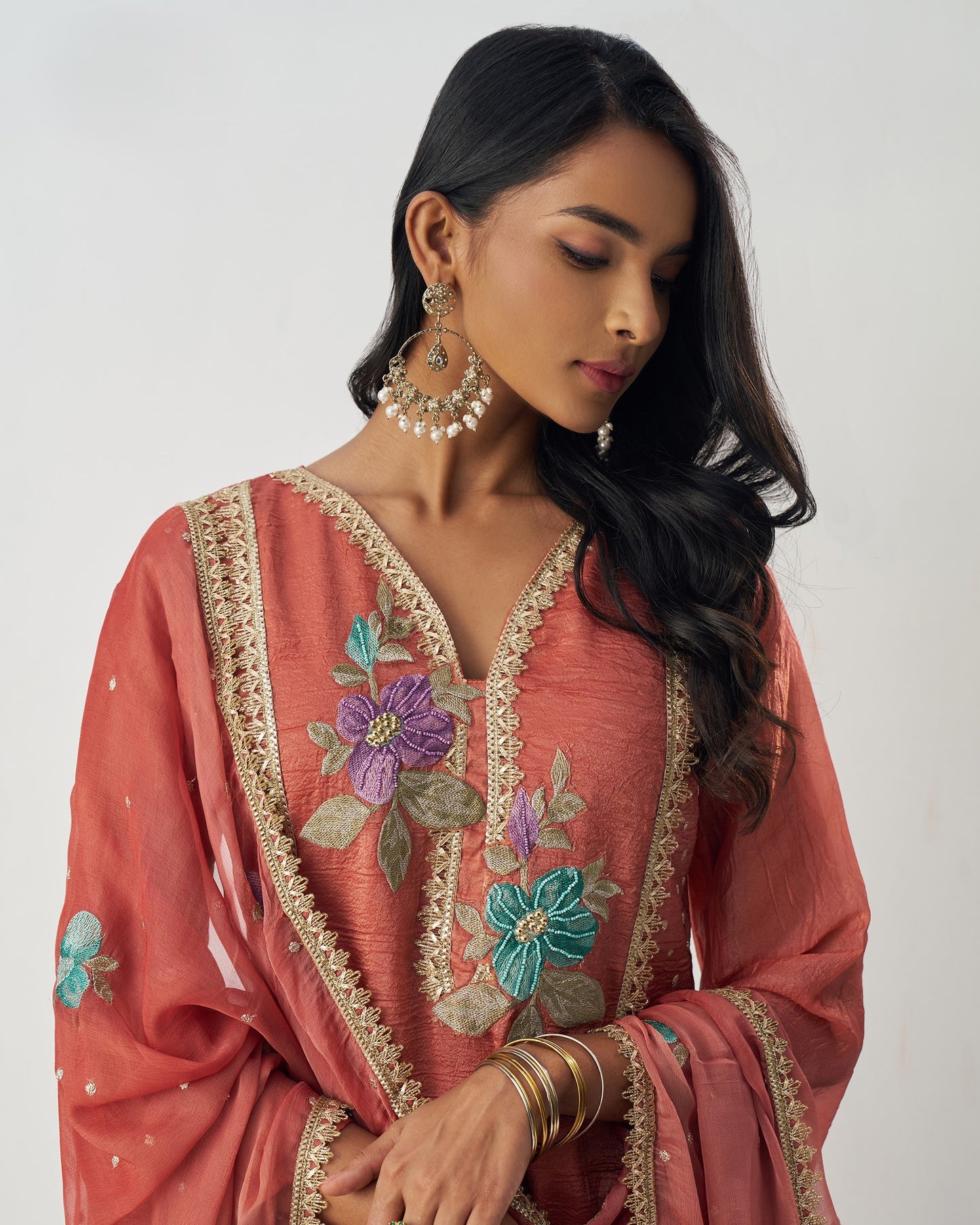 Woman wearing a coral pink embroidered traditional outfit with floral patterns on a plain background