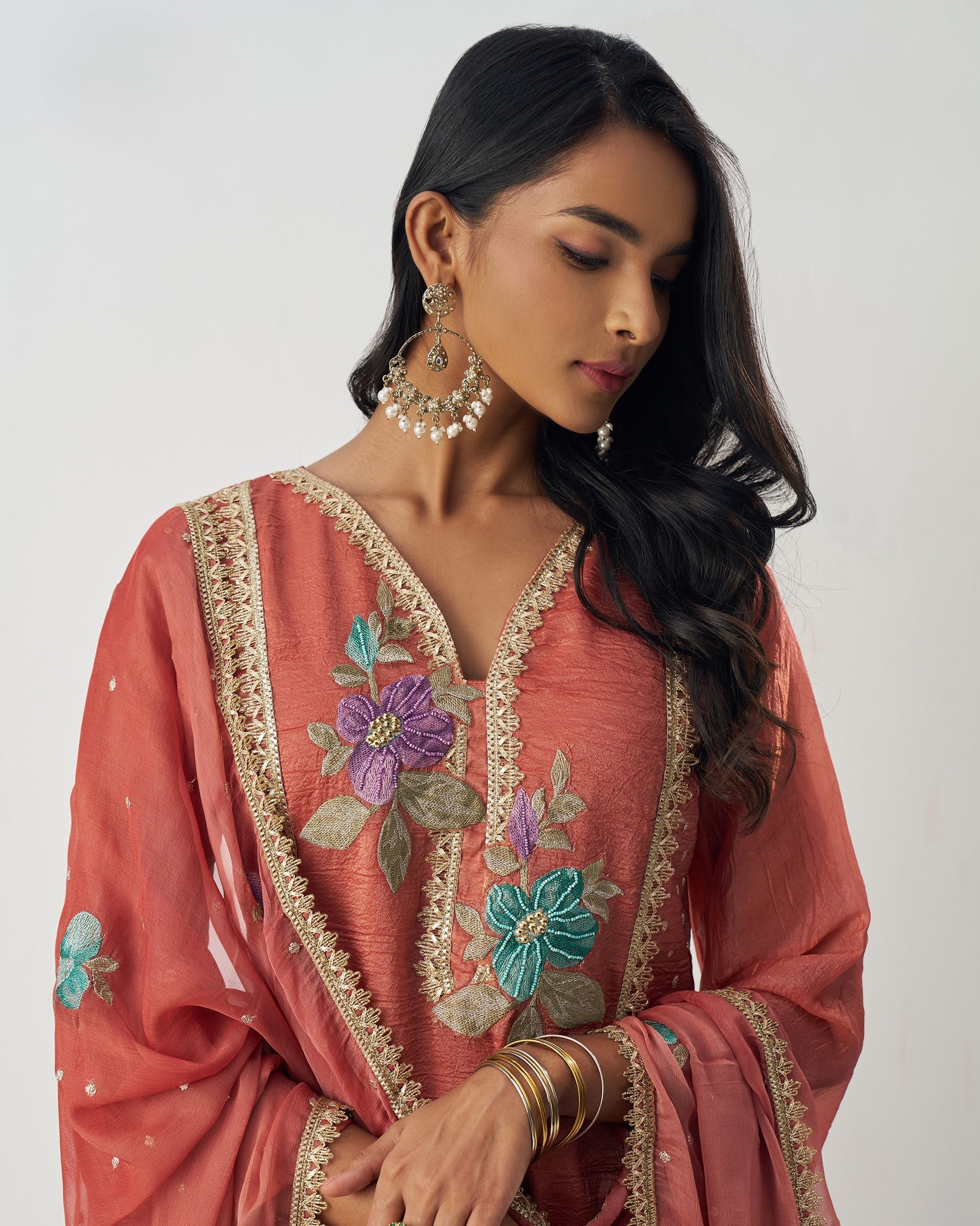 Woman wearing a coral pink embroidered traditional outfit with floral patterns on a plain background