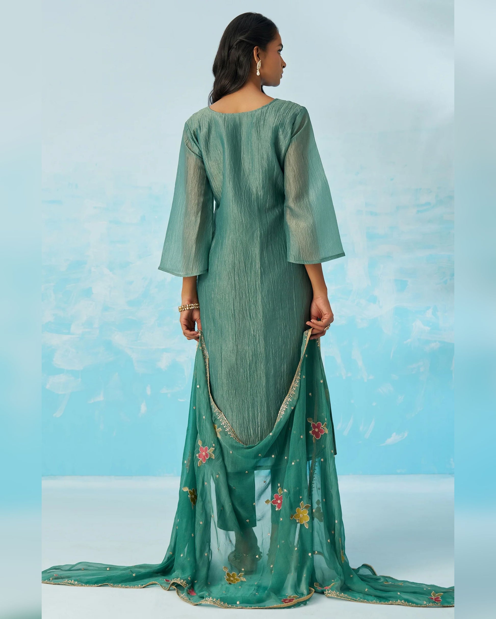 Woman wearing a green traditional outfit with a matching dupatta against a light blue background