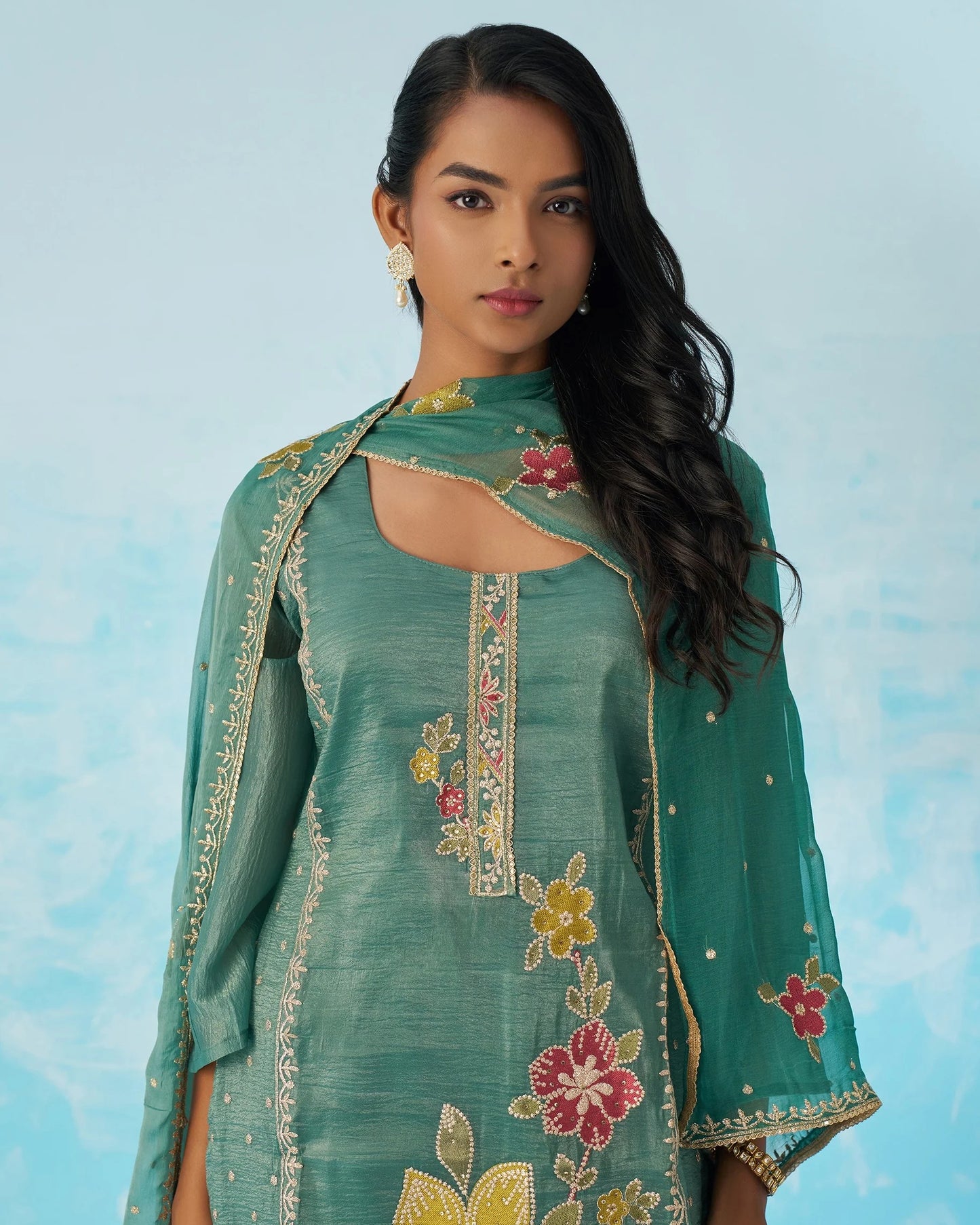 Woman wearing a teal embroidered traditional outfit against a light blue background