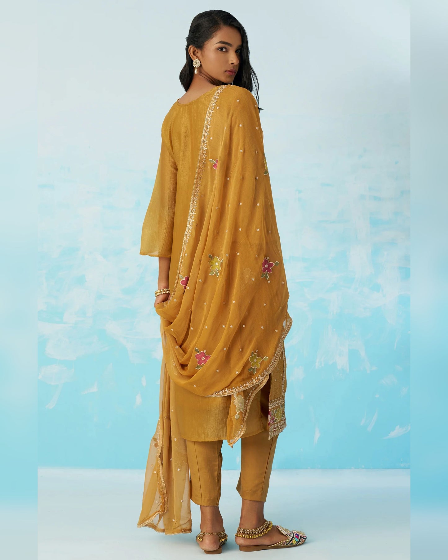 Woman wearing a mustard yellow traditional outfit with floral embroidery on a light blue background