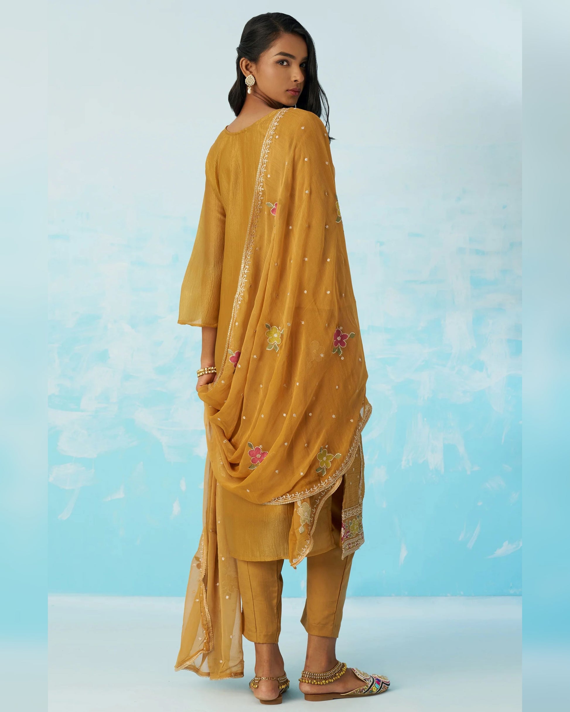 Woman wearing a mustard yellow traditional outfit with floral embroidery on a light blue background