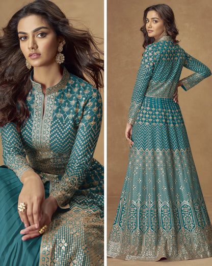 Woman wearing a teal and gold embroidered traditional outfit, sitting and standing against a beige background.
