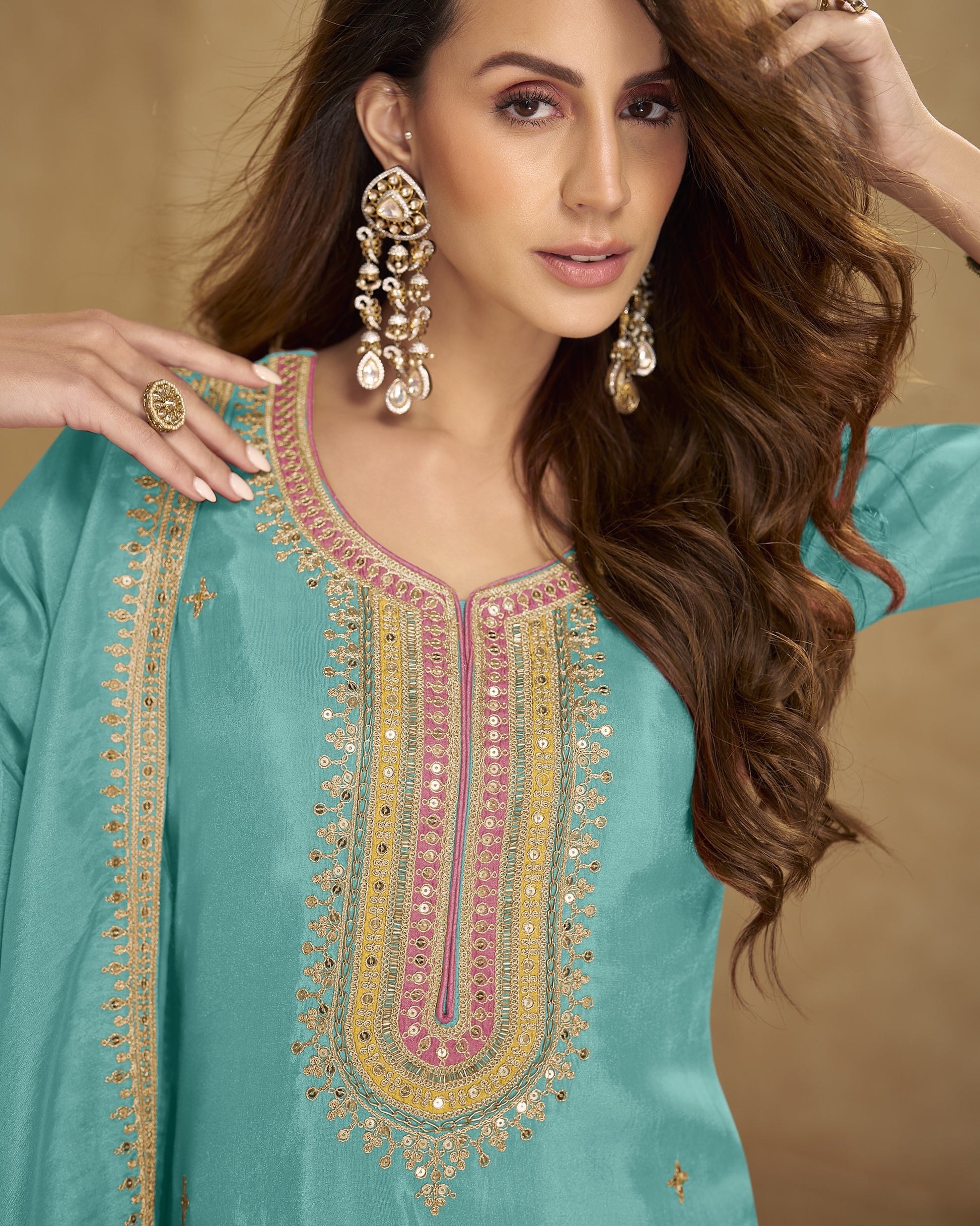 Woman wearing a turquoise embroidered dress with gold and pink details against a beige background