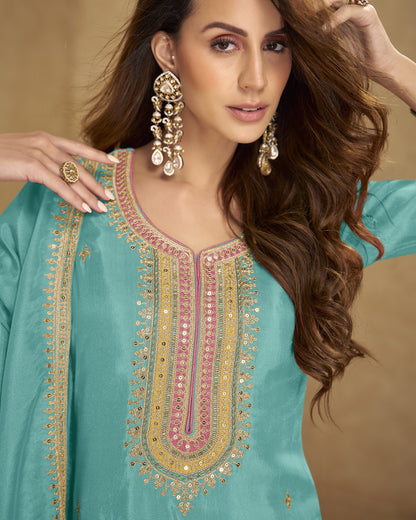 Woman wearing a turquoise embroidered dress with gold and pink details against a beige background