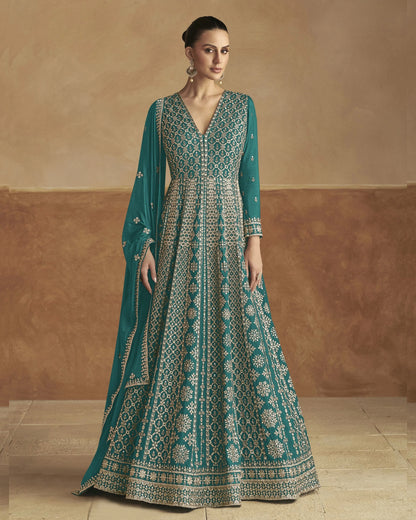 Woman wearing a teal embroidered dress against a beige wall.