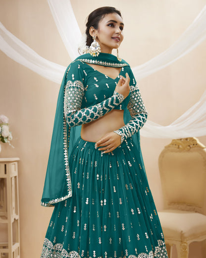 Woman wearing a teal traditional outfit with silver embroidery in a decorated room.