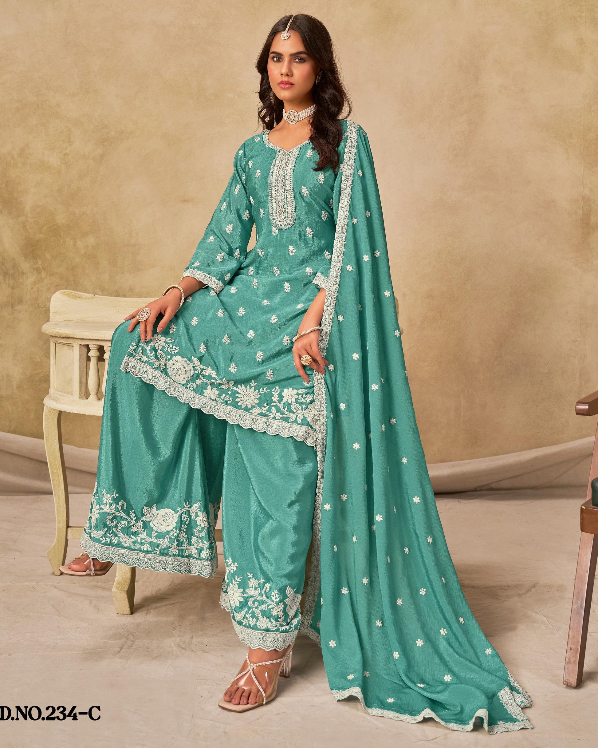 Woman wearing a teal traditional outfit with white embroidery against a beige wall.