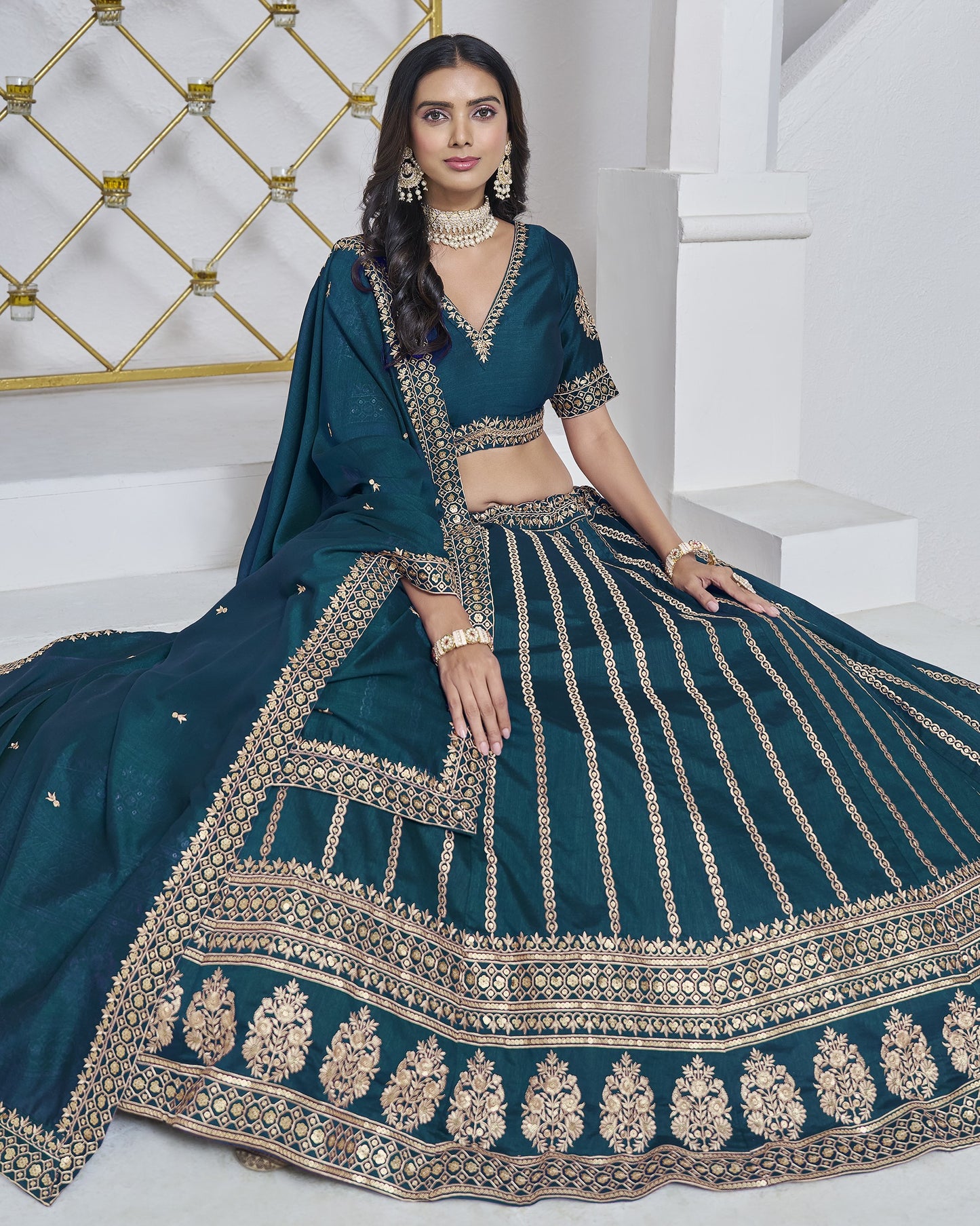 Woman wearing a teal traditional outfit with intricate gold embroidery in a white room.