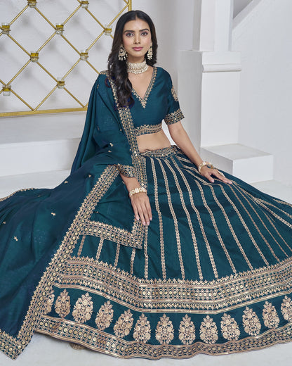 Woman wearing a teal traditional outfit with intricate gold embroidery in a white room.