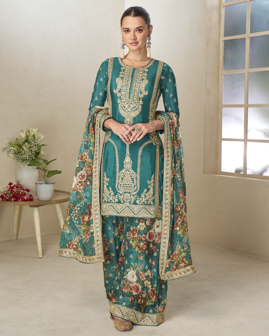 Woman wearing a teal embroidered traditional outfit in a room with a window and small table.