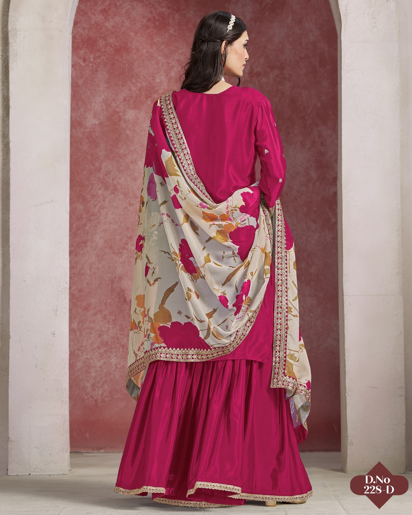 Woman wearing a pink traditional outfit with a floral shawl against a red and beige wall.