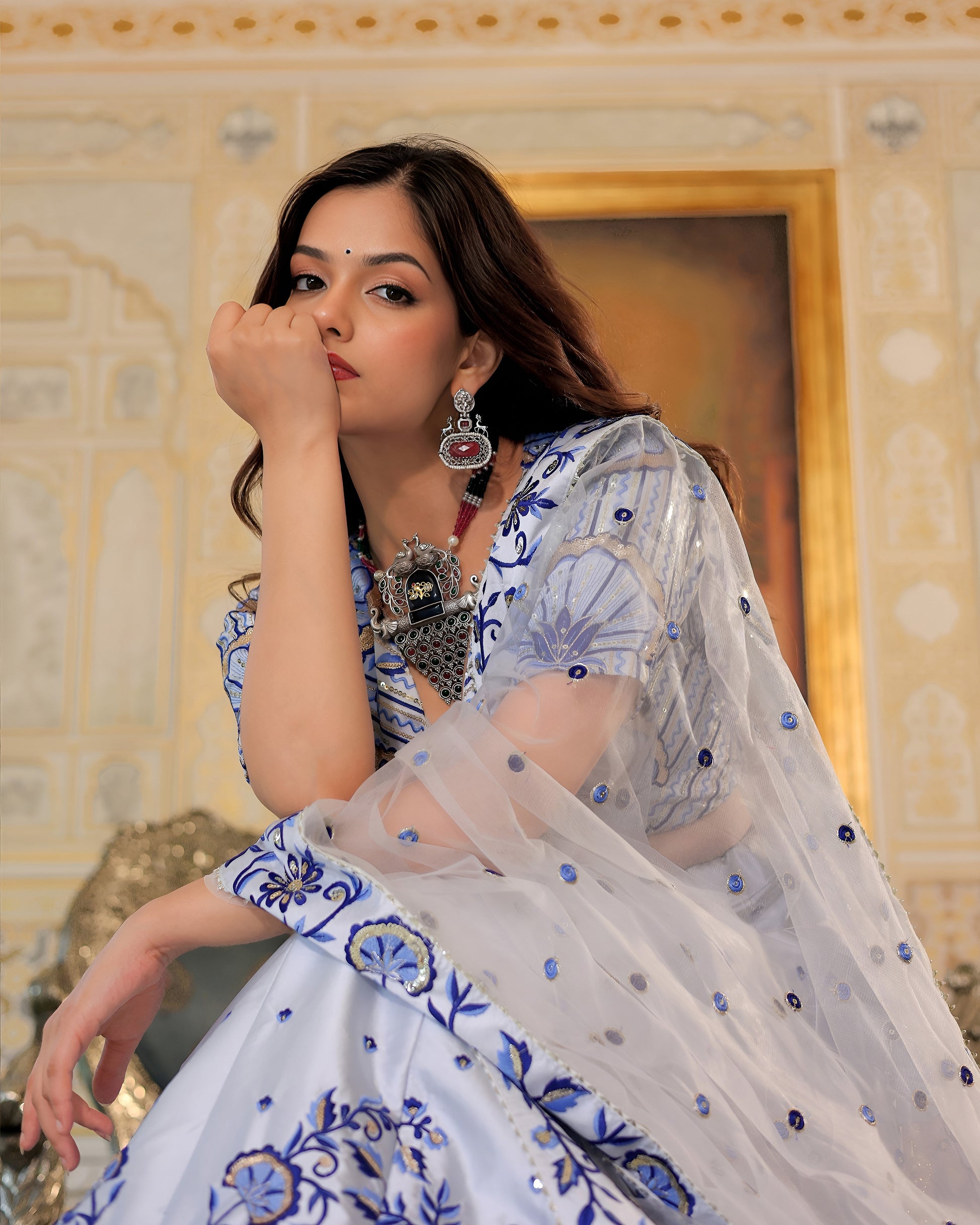 Woman in traditional attire with intricate patterns, sitting indoors.
