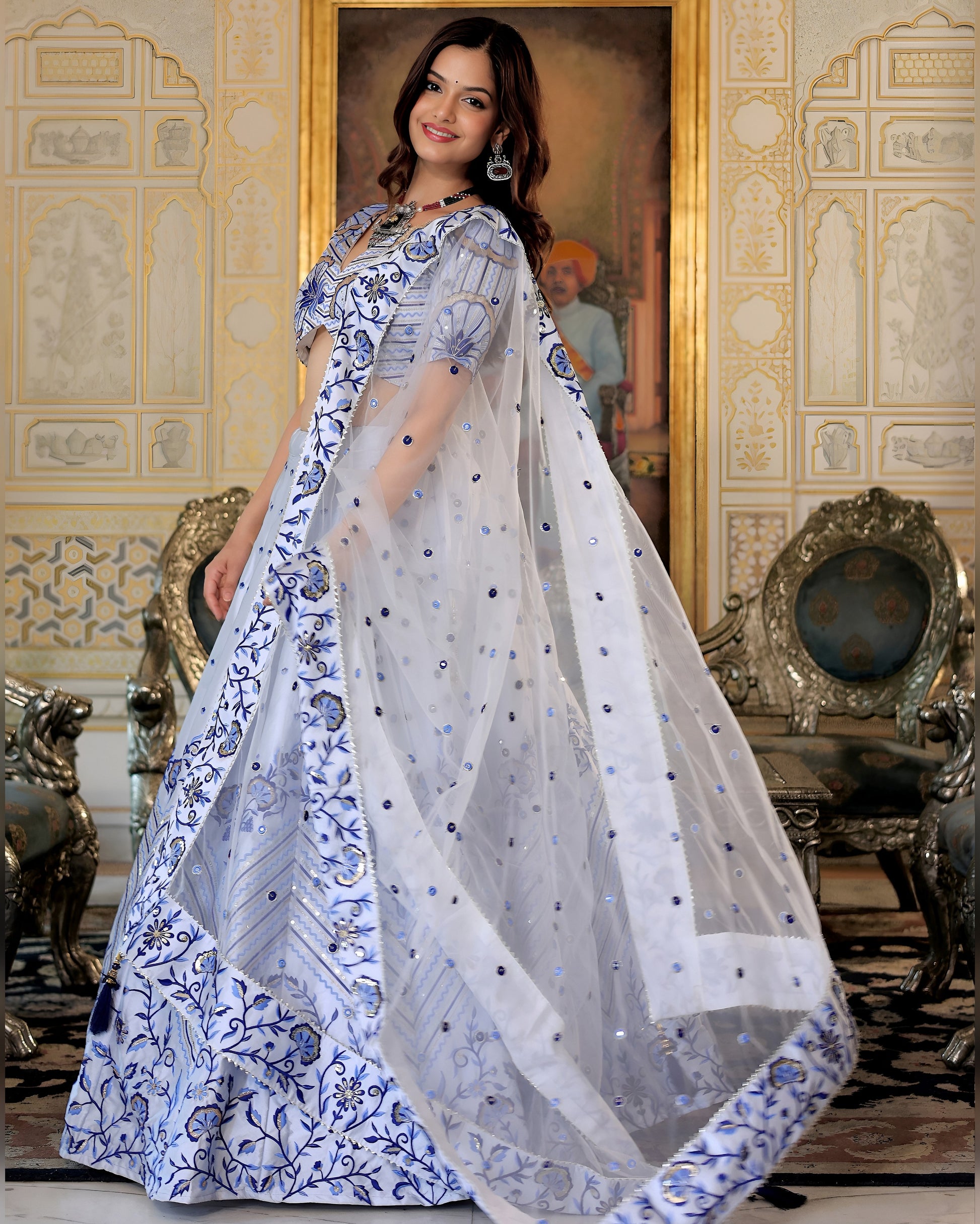 Woman wearing a white saree with blue floral patterns in an ornate room.