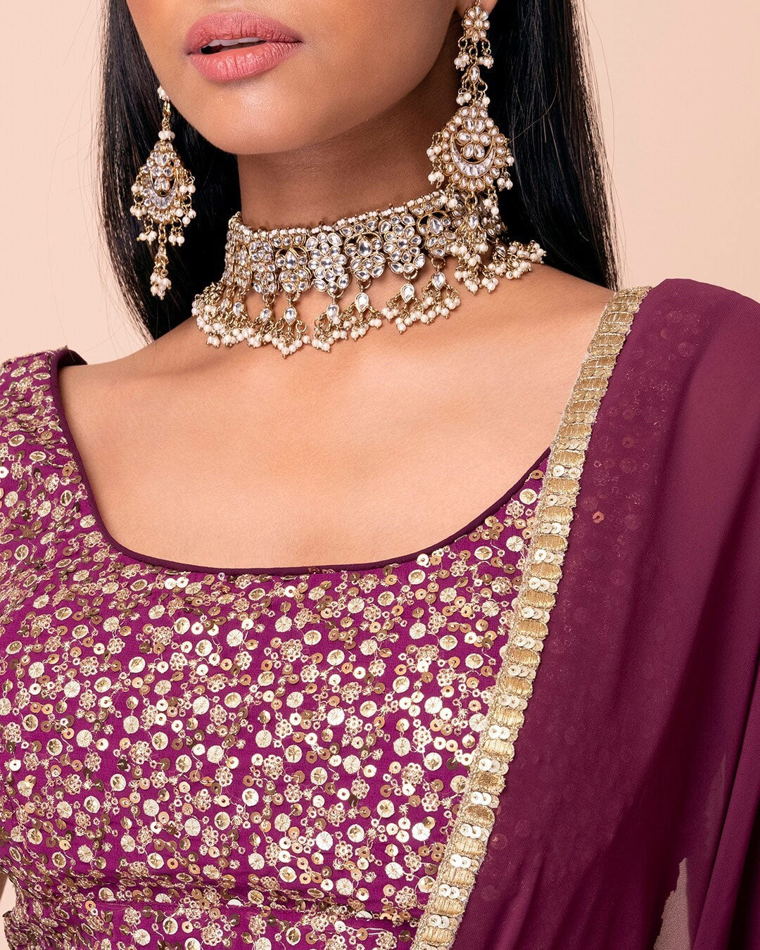 Close-up of a person wearing a purple embroidered outfit with gold jewelry on a beige background