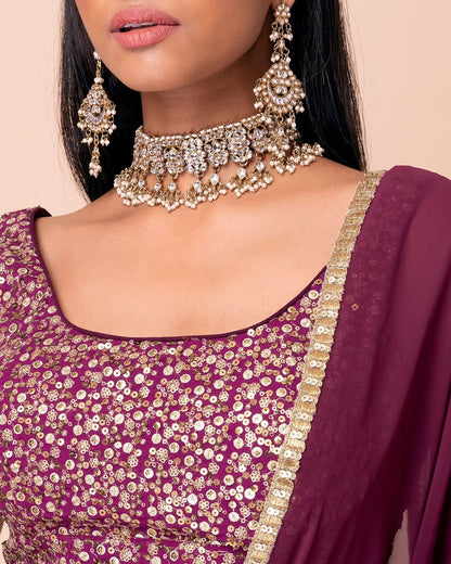 Close-up of a person wearing a purple embroidered outfit with gold jewelry on a beige background