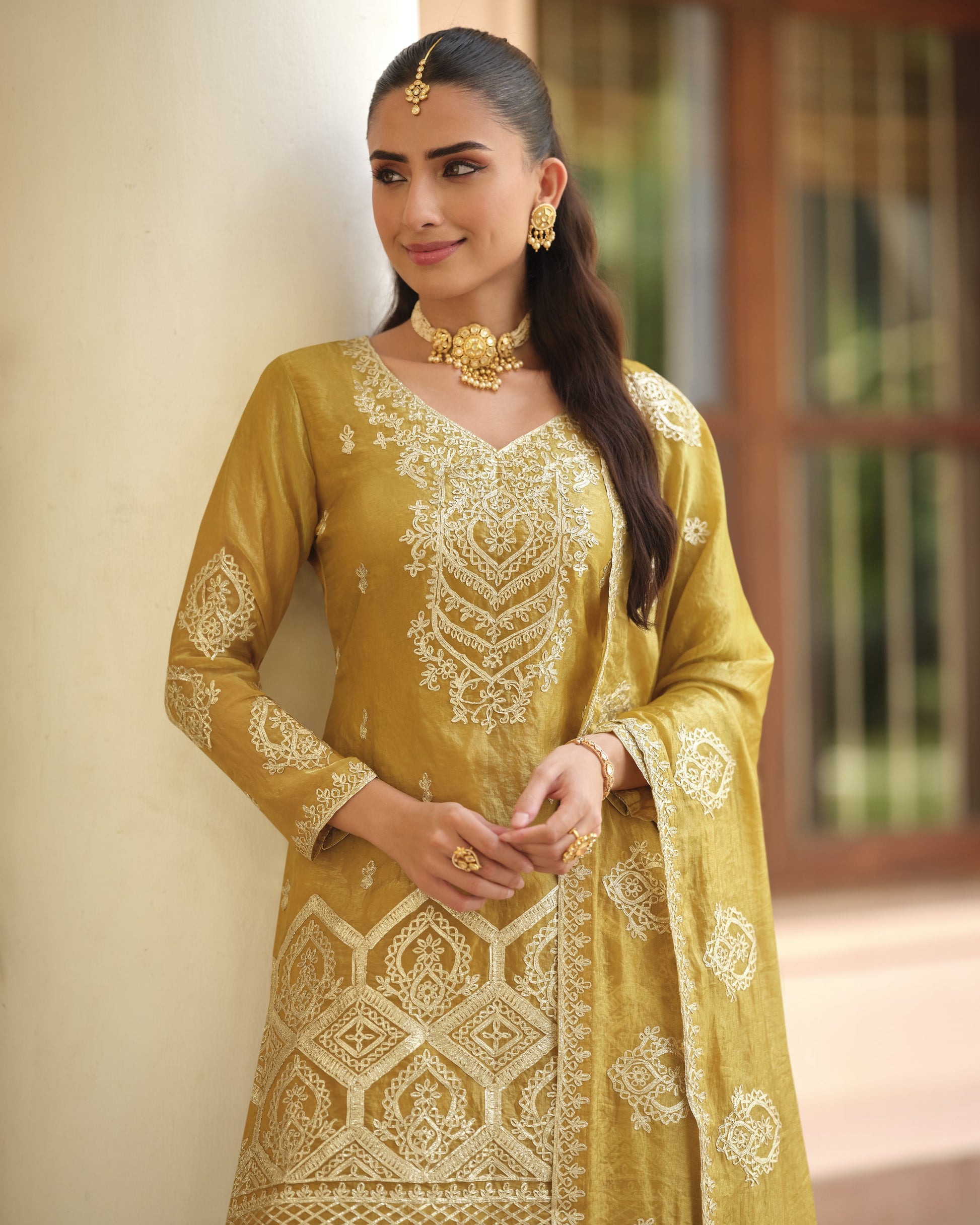 Woman wearing a yellow embroidered traditional outfit with gold jewelry against a blurred indoor background.