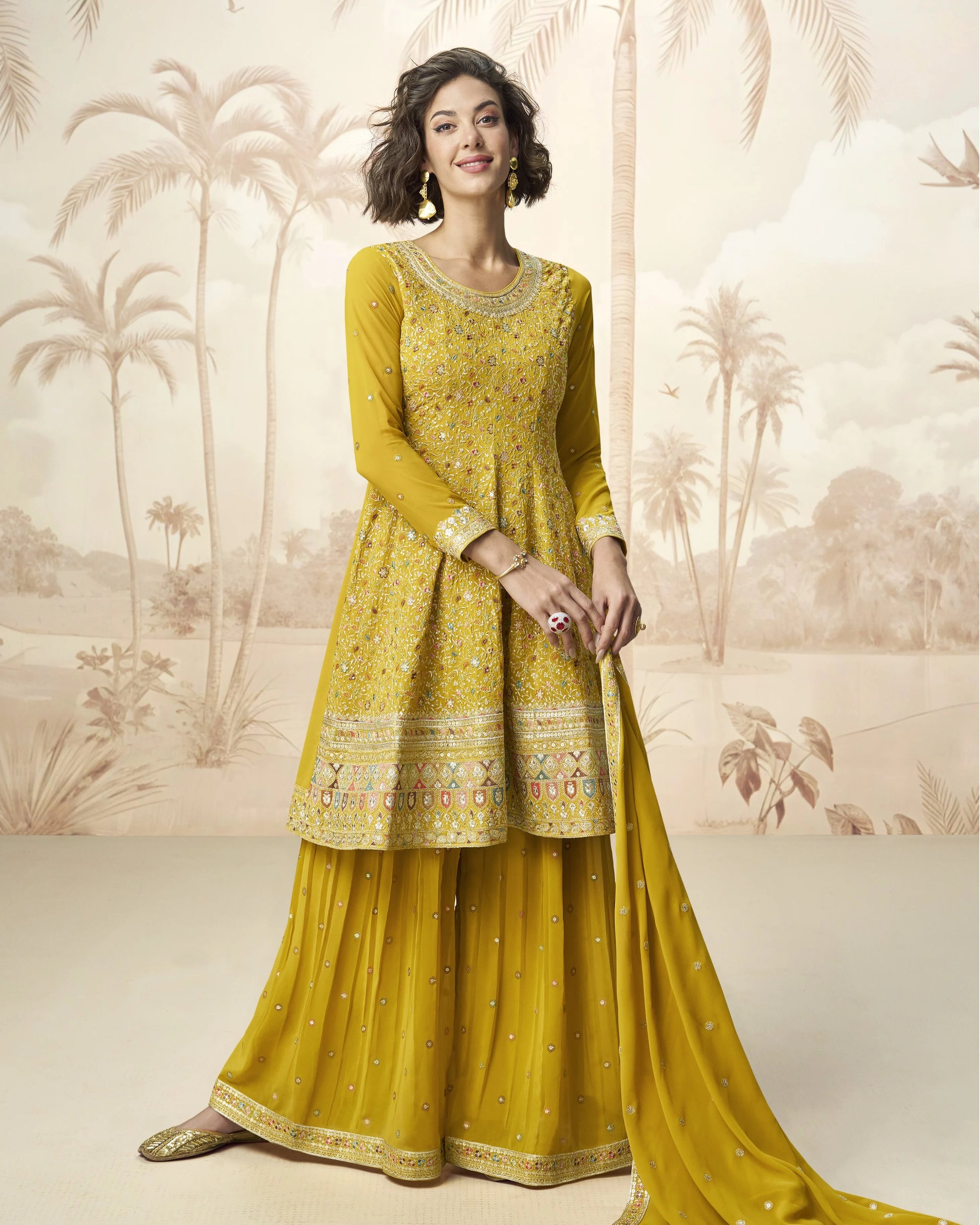 Woman wearing a yellow traditional outfit with intricate patterns against a decorative wall.