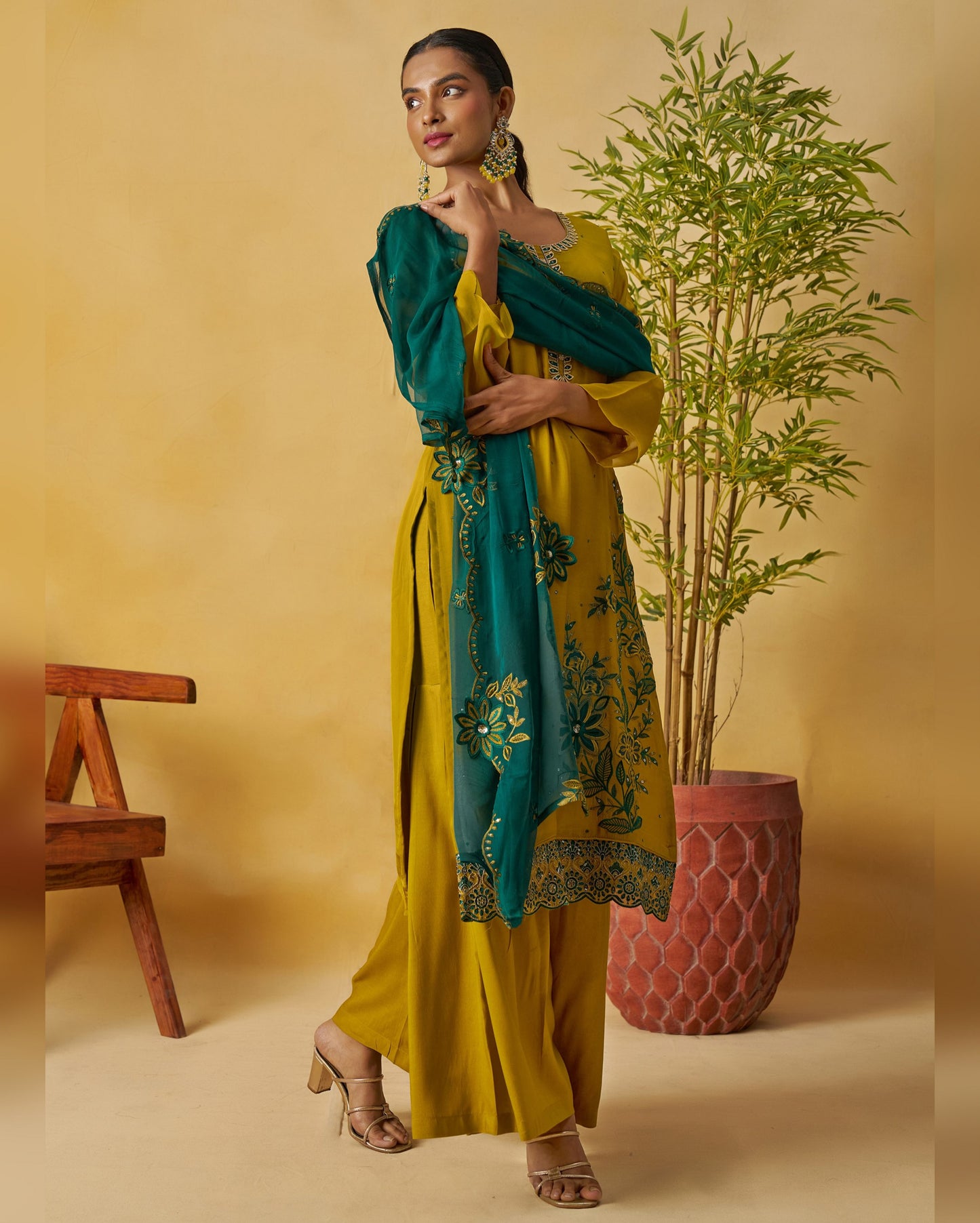 Woman in a yellow and teal traditional outfit with a plant and wooden bench in the background