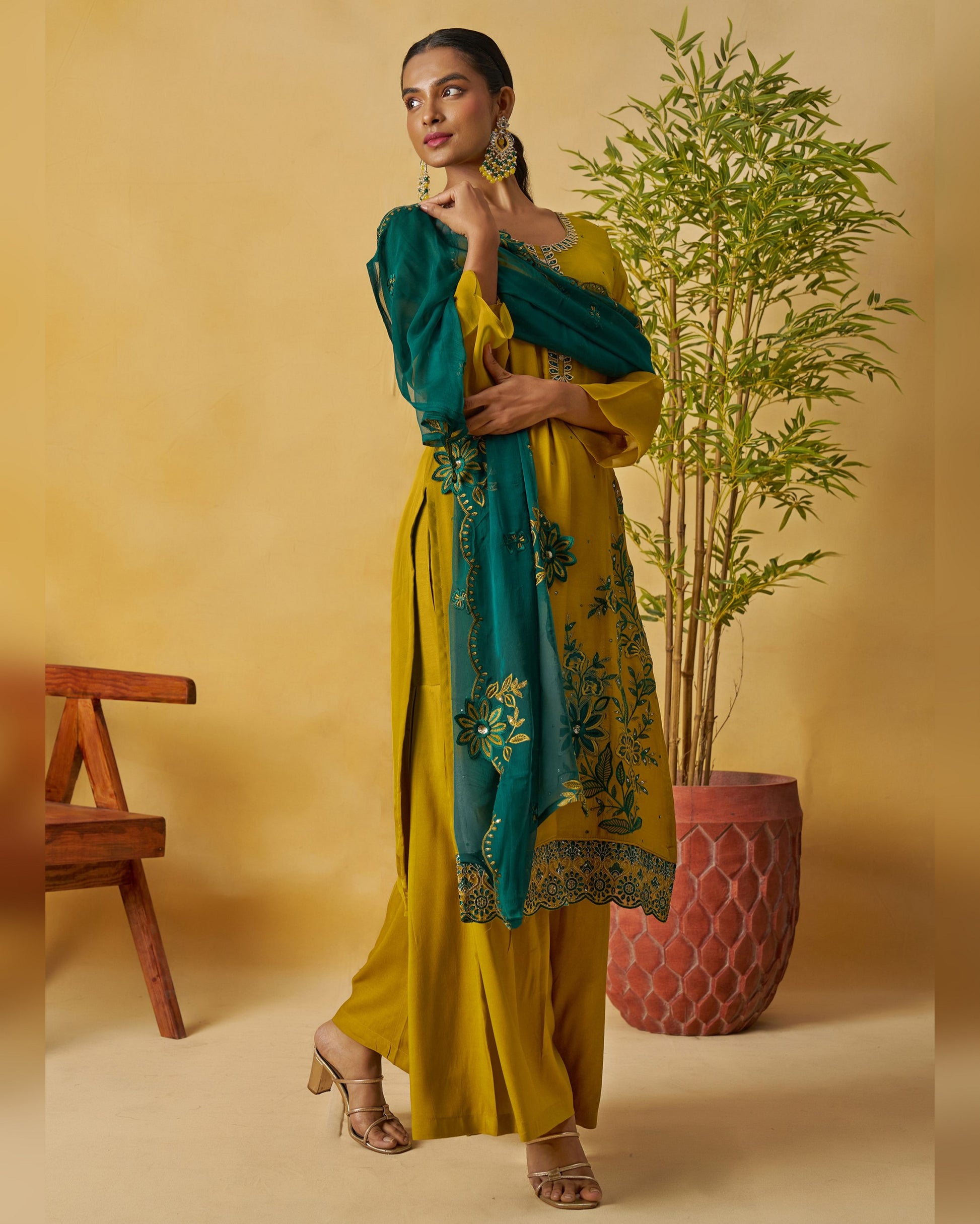 Woman in a yellow and teal traditional outfit with a plant and wooden bench in the background