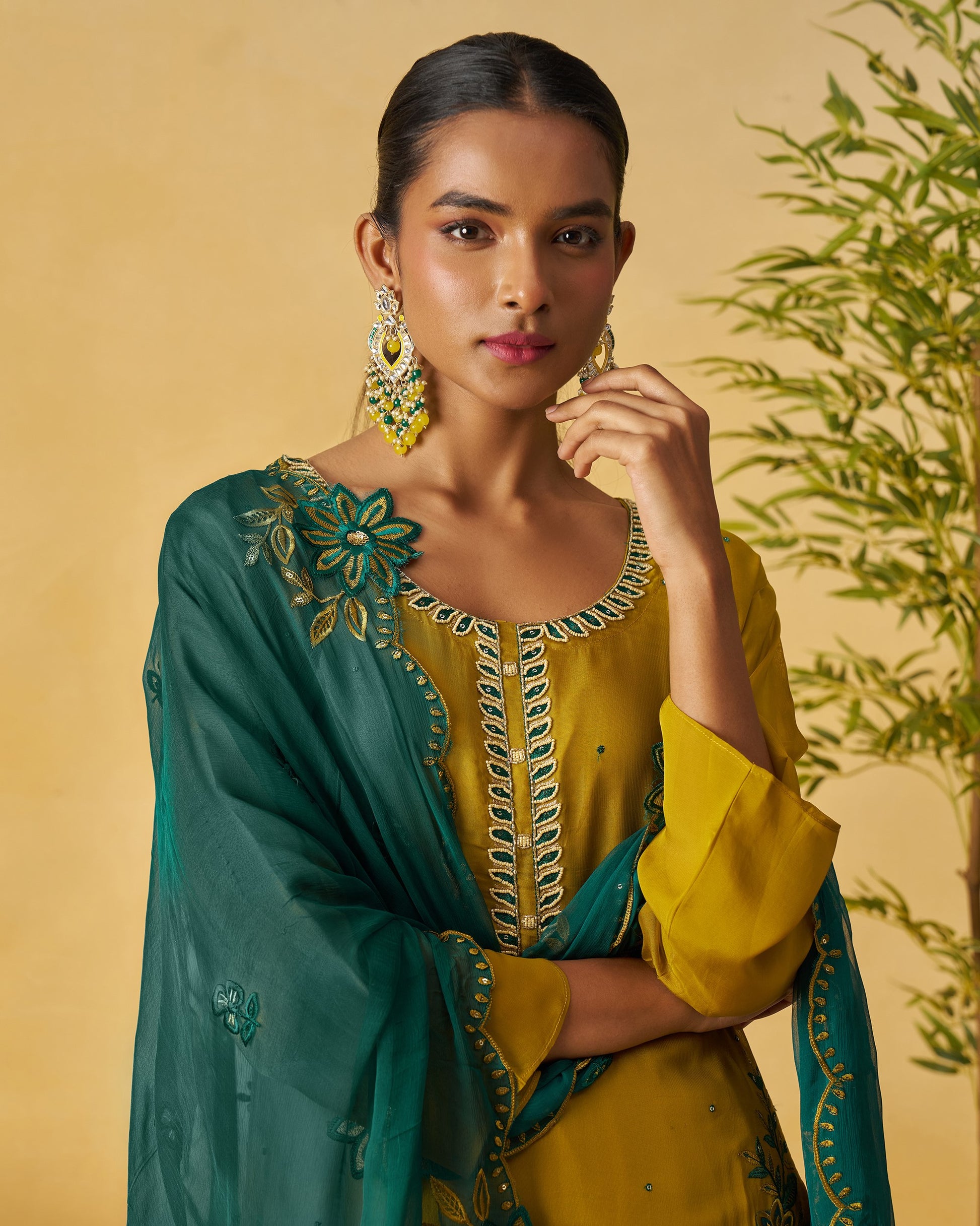 Woman wearing a yellow and green traditional outfit with floral embroidery against a beige background.