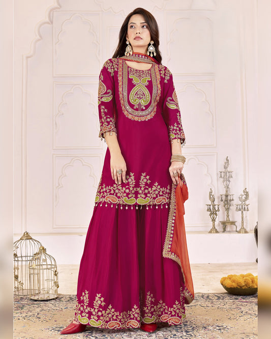 Woman wearing a vibrant pink and orange traditional outfit with intricate embroidery in a decorative setting.