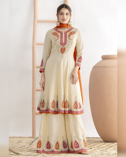 Woman wearing a cream-colored traditional outfit with orange and red embroidery in a minimalistic setting.
