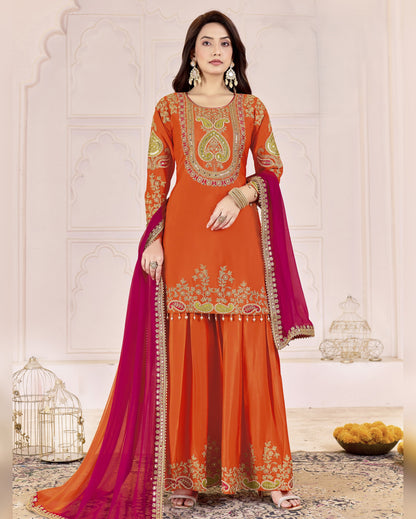 Woman wearing an orange and pink traditional outfit with intricate embroidery in a decorative setting.