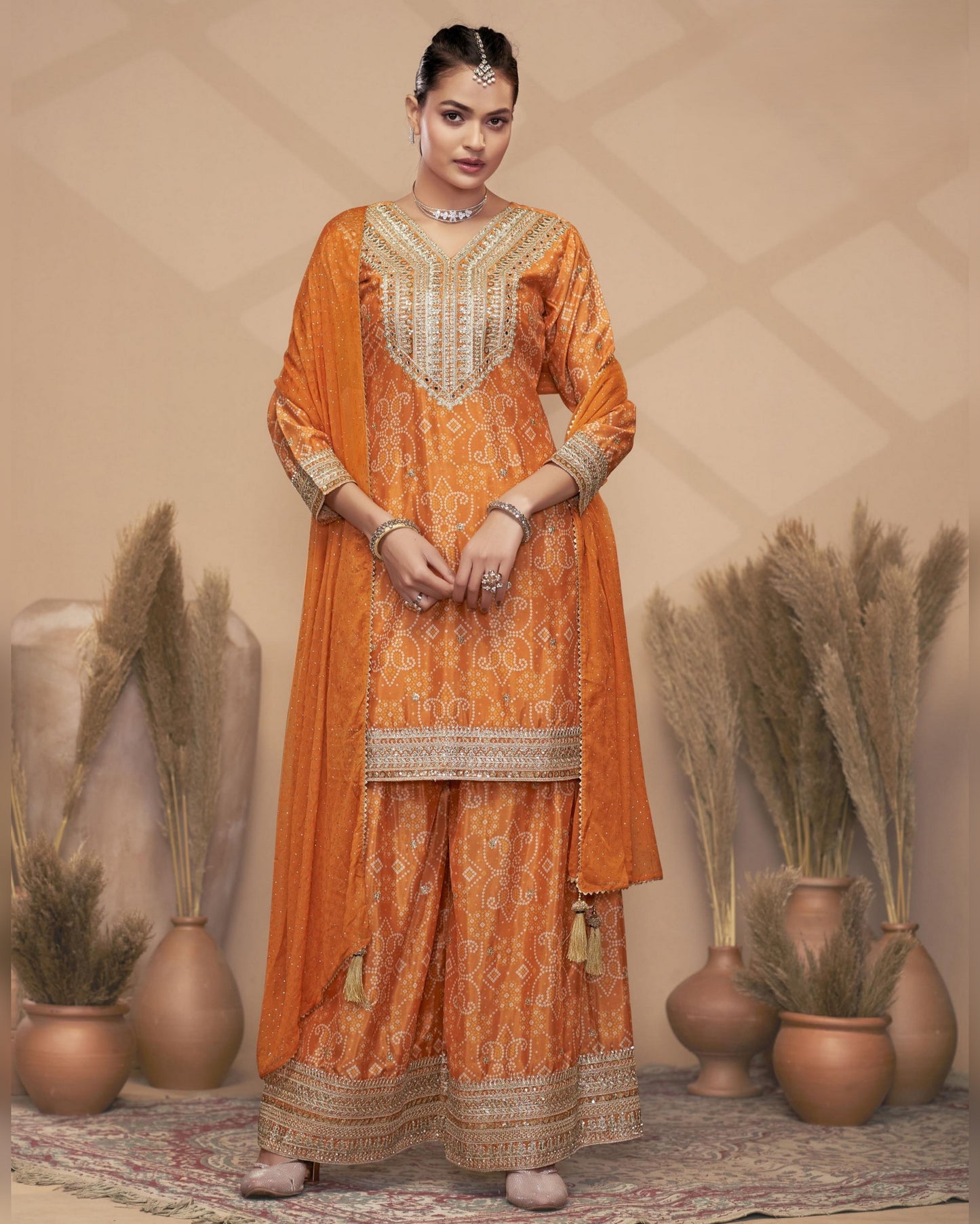 Woman in an orange embroidered traditional outfit standing against a beige wall with decorative plants.