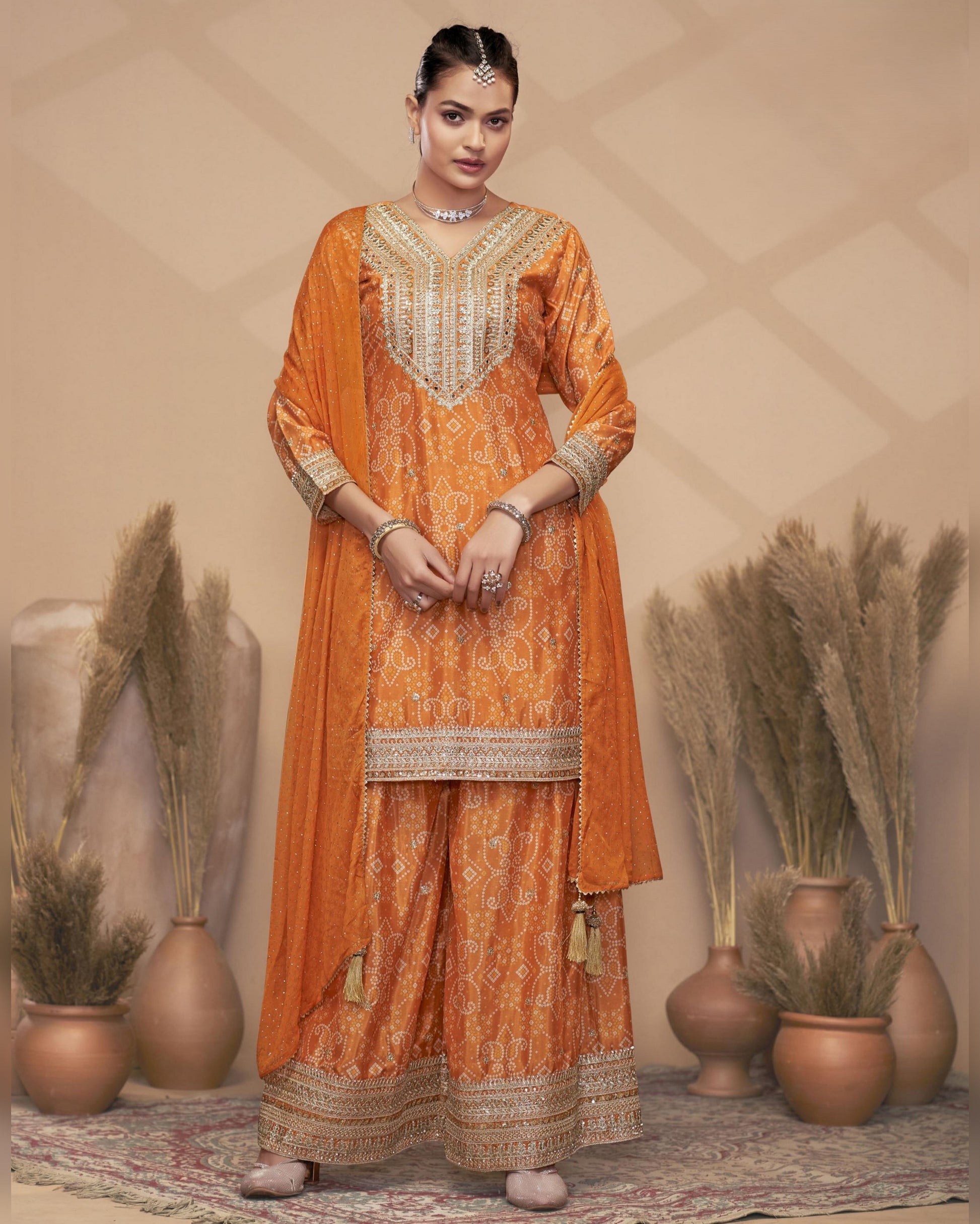 Woman in an orange embroidered traditional outfit standing against a beige wall with decorative plants.