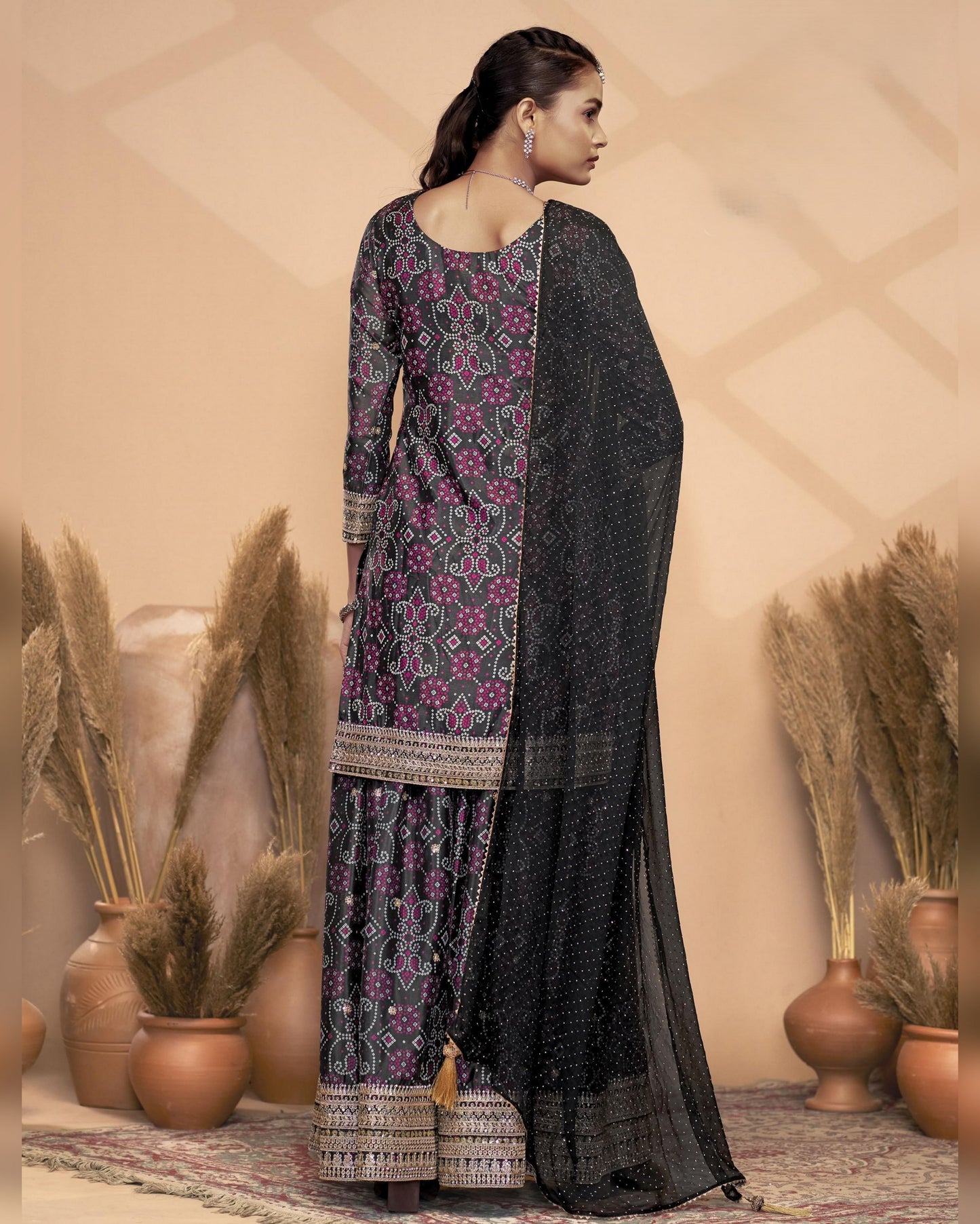 Woman wearing a black and purple patterned traditional outfit with a beige wall and decorative plants in the background.