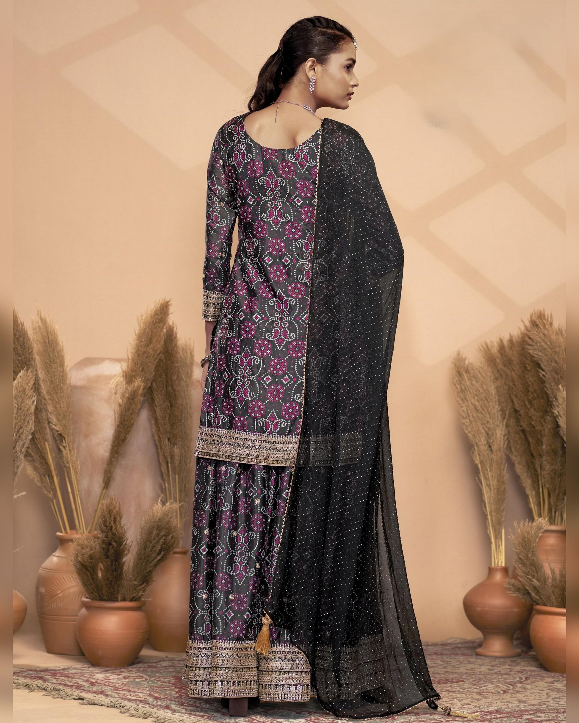 Woman wearing a black and purple patterned traditional outfit with a beige wall and decorative plants in the background.