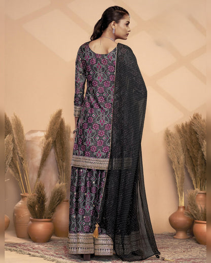 Woman wearing a black and purple patterned traditional outfit with a beige wall and decorative plants in the background.