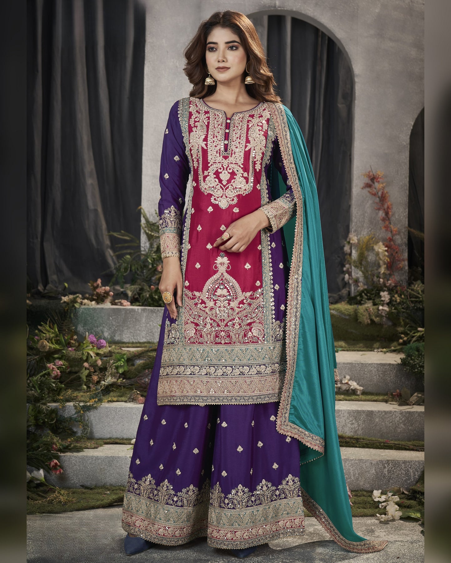 Woman wearing a traditional embroidered outfit with a purple and pink design, standing in an outdoor setting.