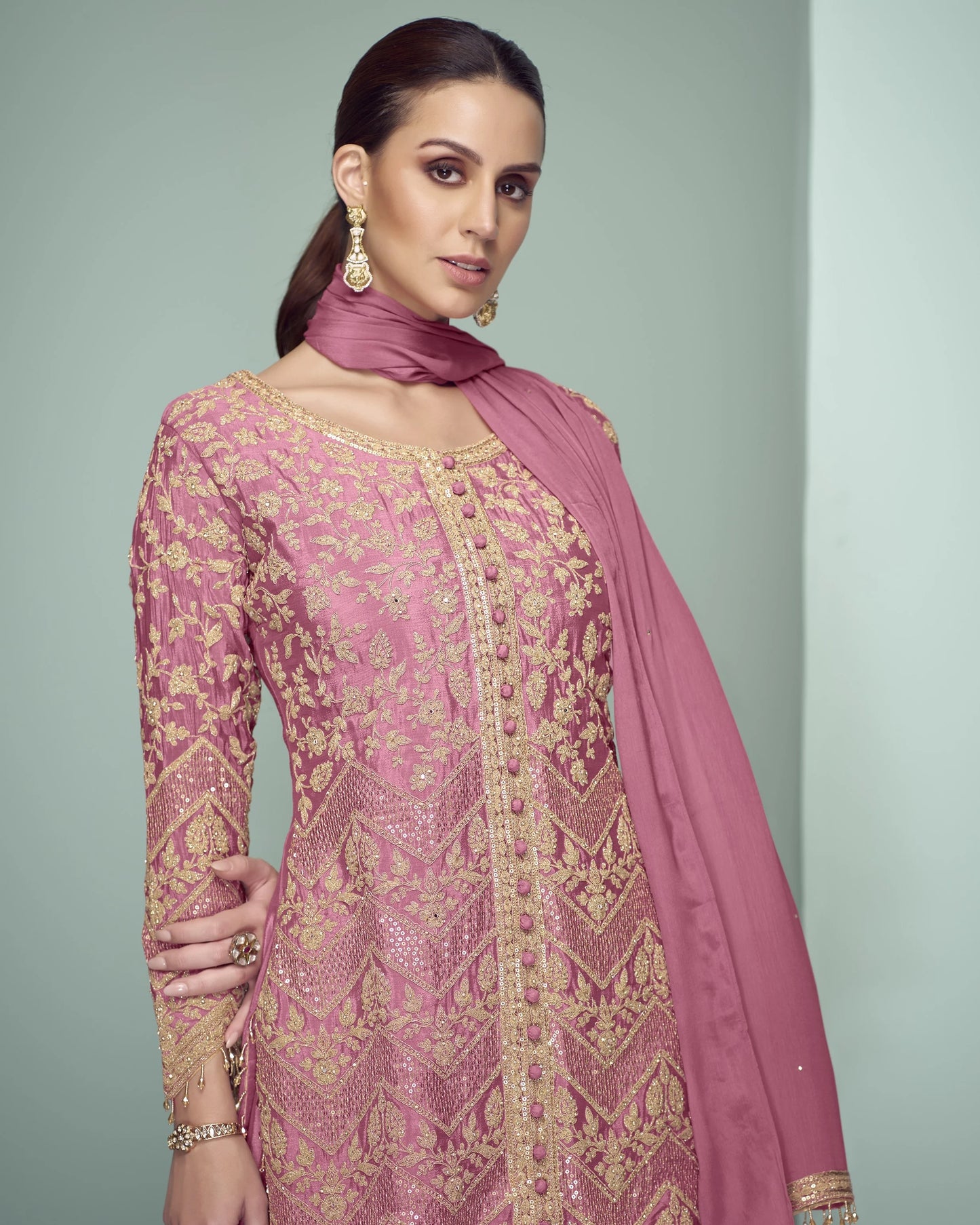 Woman wearing a pink embroidered traditional outfit with a matching dupatta against a gray background